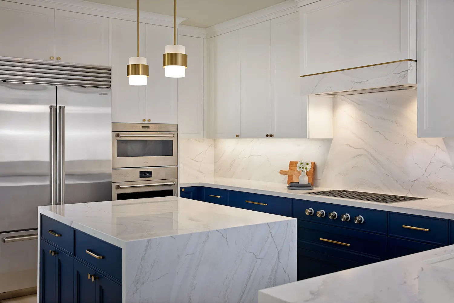 Navy and white kitchen with a stainless refrigerator, marble waterfall island and brass pendants