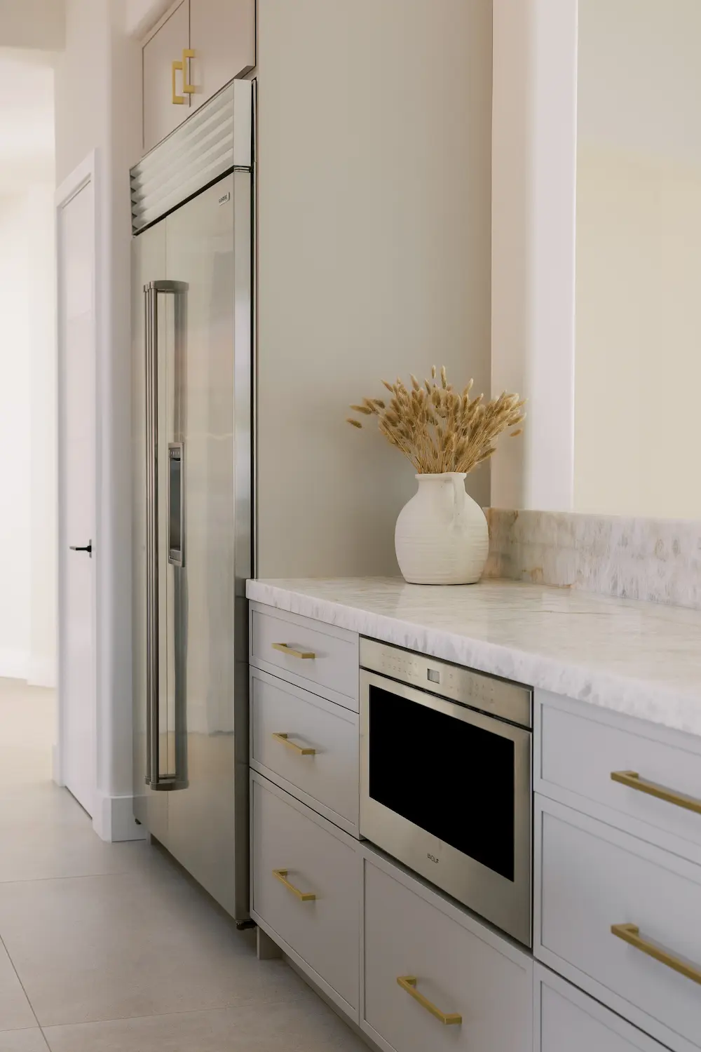 Stainless built-in refrigerator beside gray cabinets with brass pulls and a drawer microwave