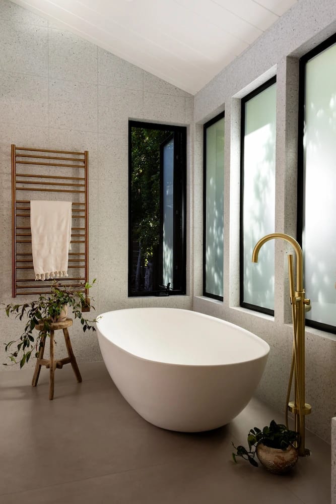 Freestanding white soaking tub beside tall windows and a brass floor-mounted filler