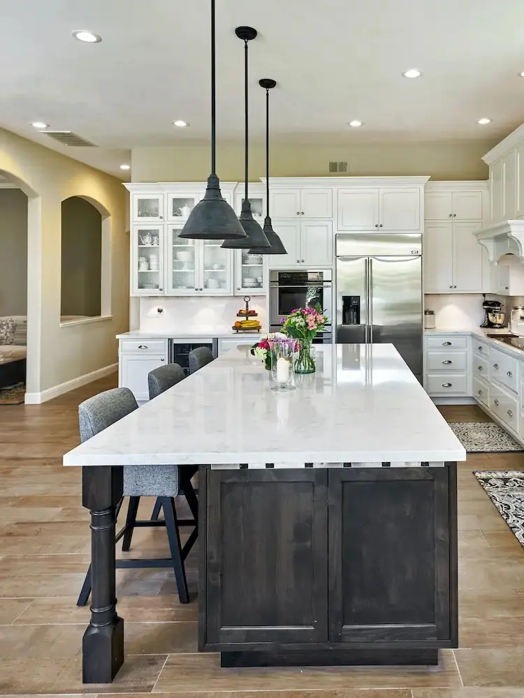 Spacious kitchen with a large white island, dark wood base, white cabinetry, stainless steel appliances, and black pendant lights, featuring decorative rugs and ample natural light