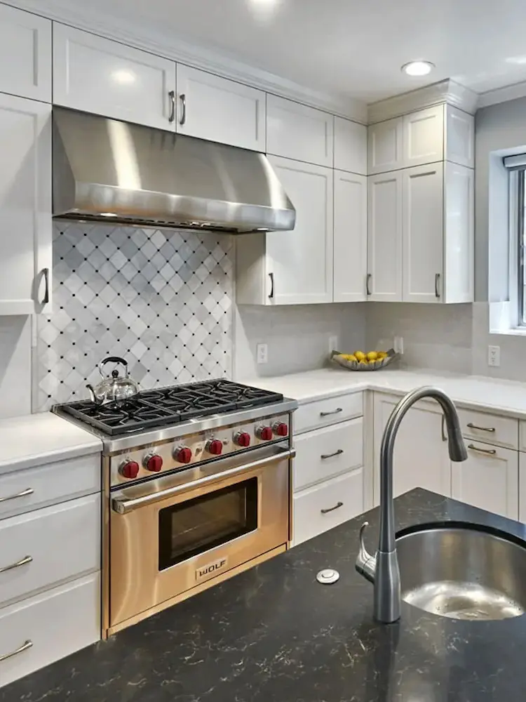 Modern kitchen featuring white cabinetry, a stainless steel Wolf range with red knobs, diamond-patterned tile backsplash, and a sleek black marble countertop with an integrated sink