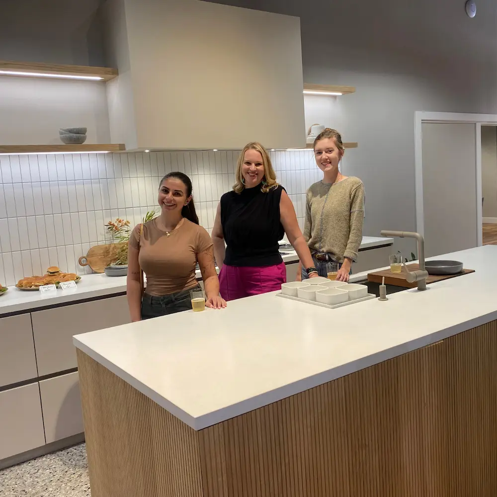 Group of designers standing in a contemporary kitchen with a sleek white countertop, wood accents, and open shelving at a social event