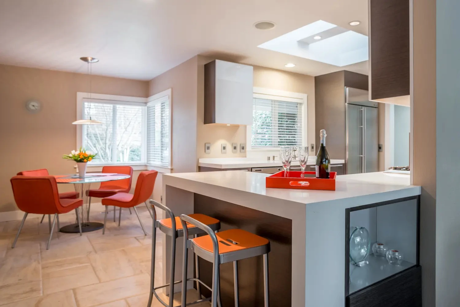 Light-filled modern kitchen with white waterfall island, orange bar stools, and open dining area with red upholstered chairs