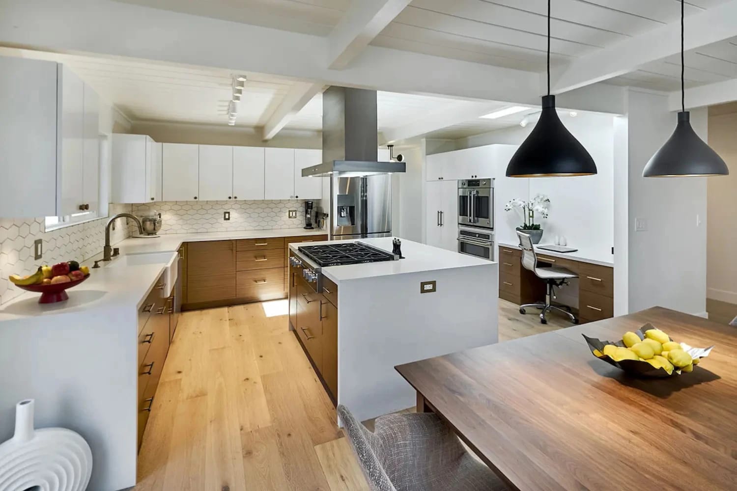 Open concept kitchen with white and wood cabinetry, hex tile backsplash, and a built-in desk nook beside double wall ovens