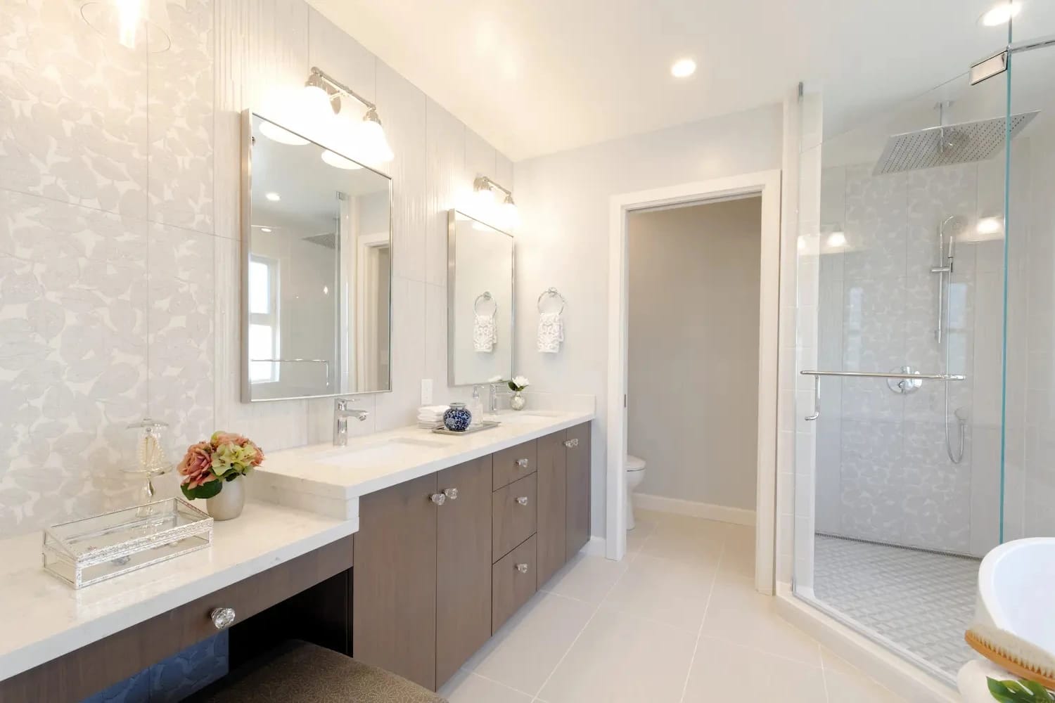 Elegant primary bathroom with double vanity, makeup counter, glass shower enclosure, and soft floral-patterned wall tile