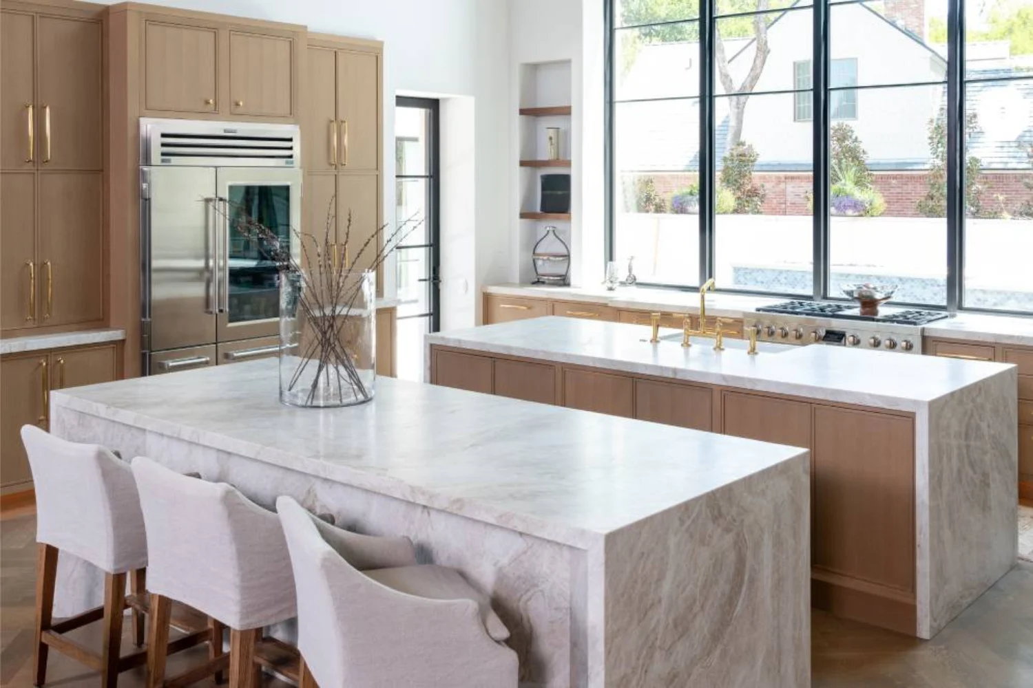 Light wood kitchen with waterfall marble island and brass accents, featuring large black-framed windows and a stainless steel fridge