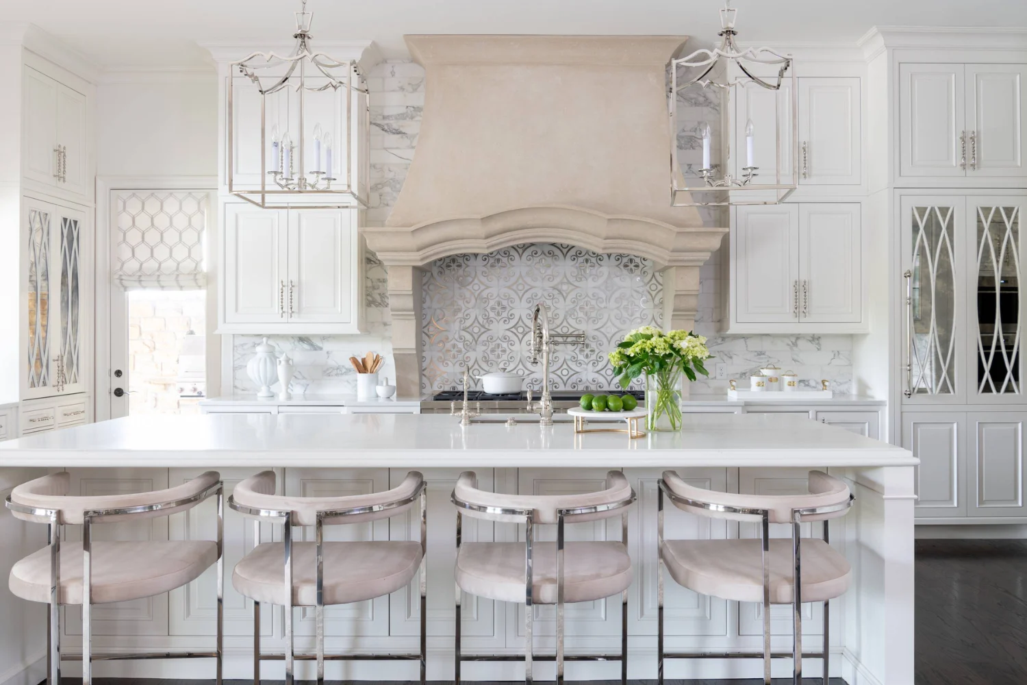 Classic white kitchen with a carved range hood, patterned tile backsplash, and elegant upholstered bar stools