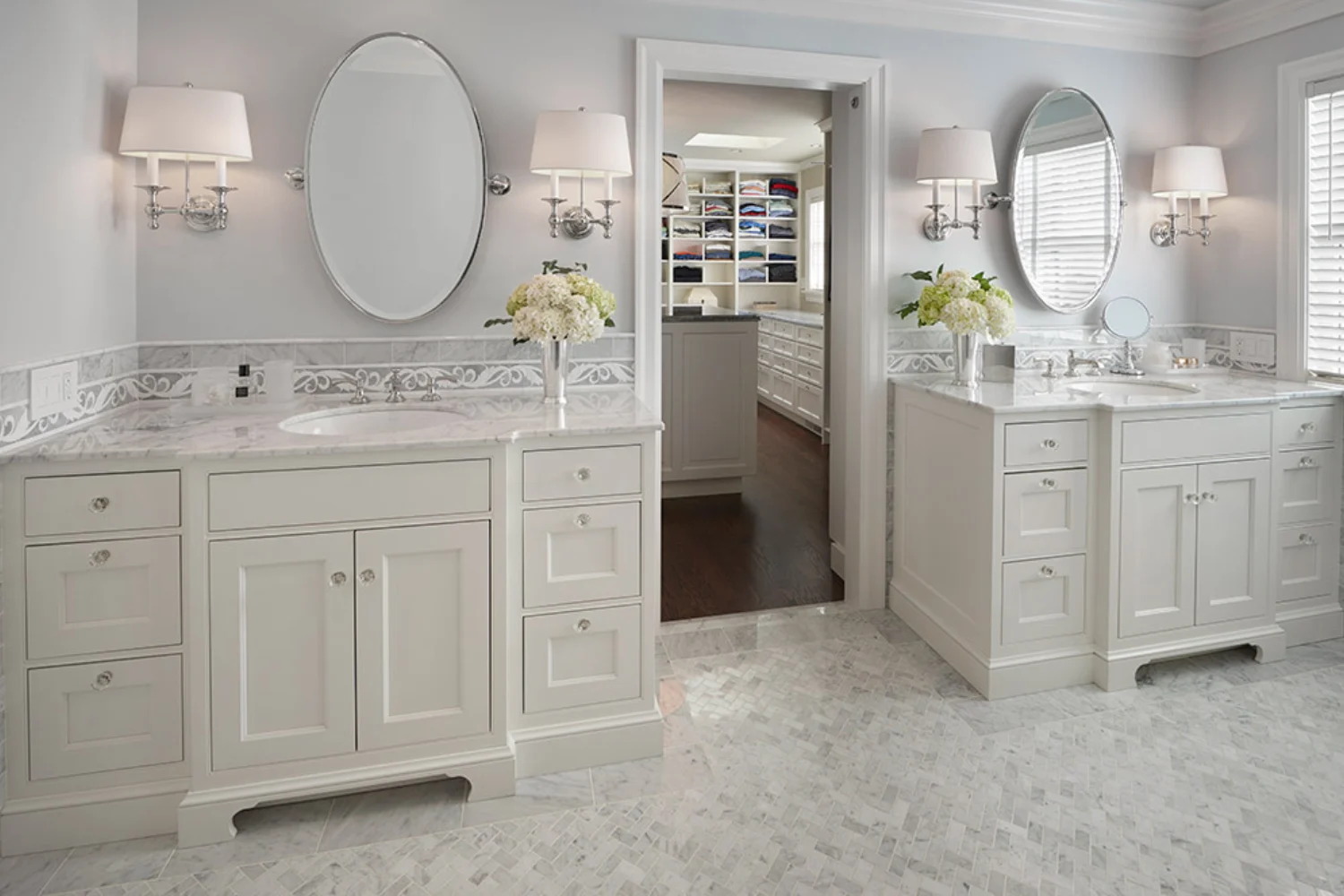 Elegant master bathroom with dual vanities, oval mirrors, white cabinetry, and a walk-in closet visible through the center doorway