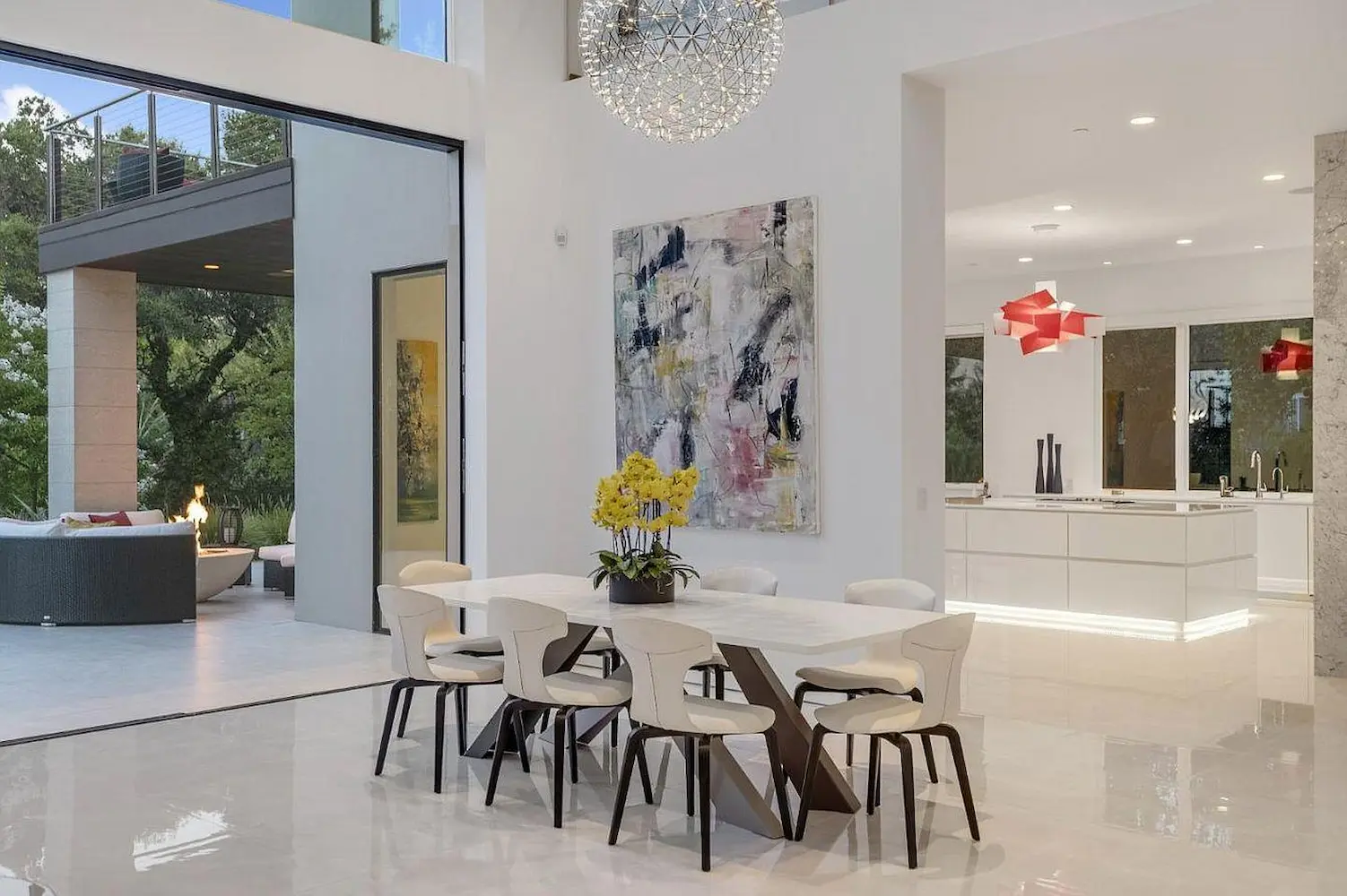Dining area with modern white chairs and a sleek rectangular table, complemented by a large abstract wall painting and a chandelier, opening to an outdoor living space