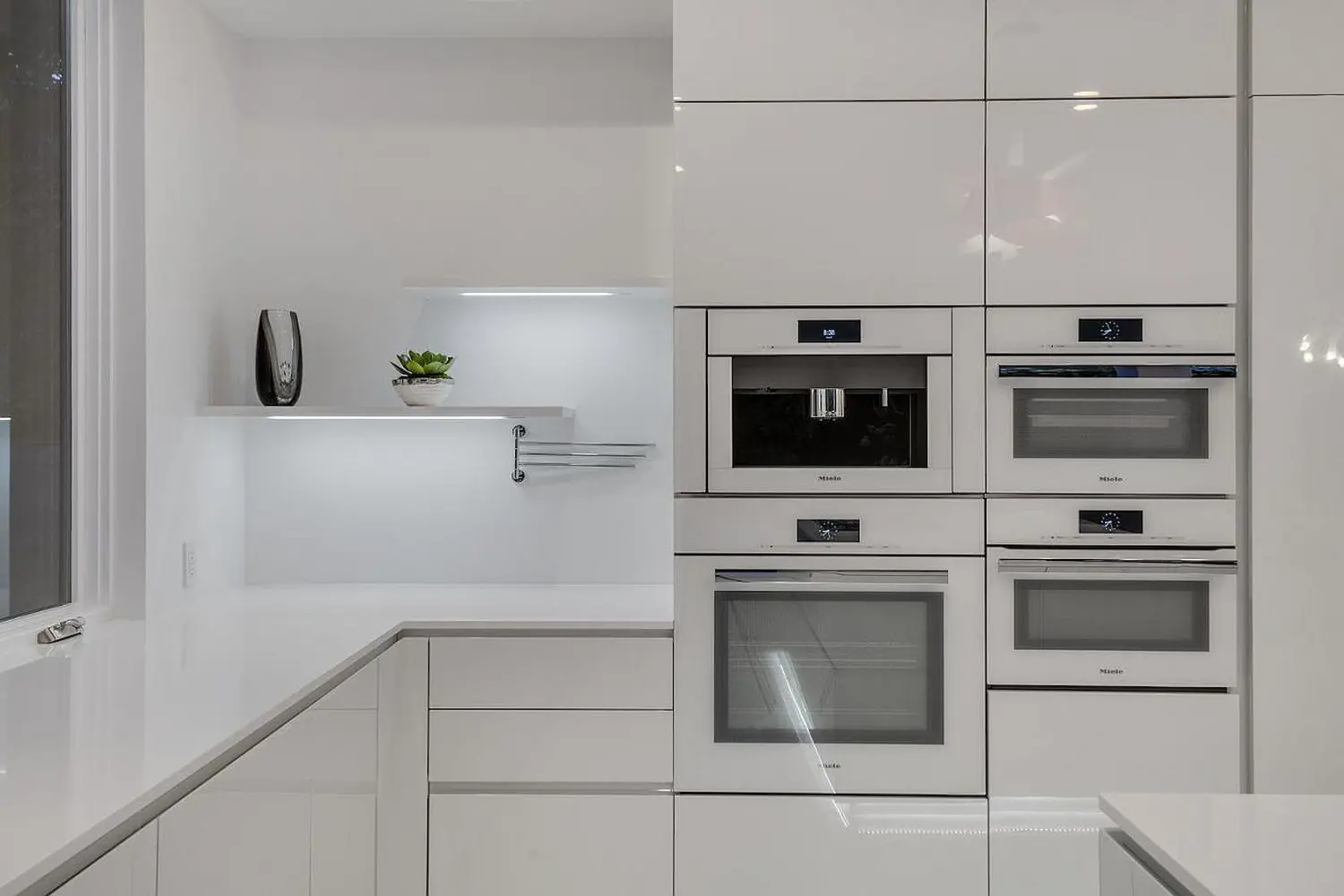 Minimalist kitchen with high-end built-in stainless steel appliances, white cabinetry, and a modern open shelving unit with decorative accents