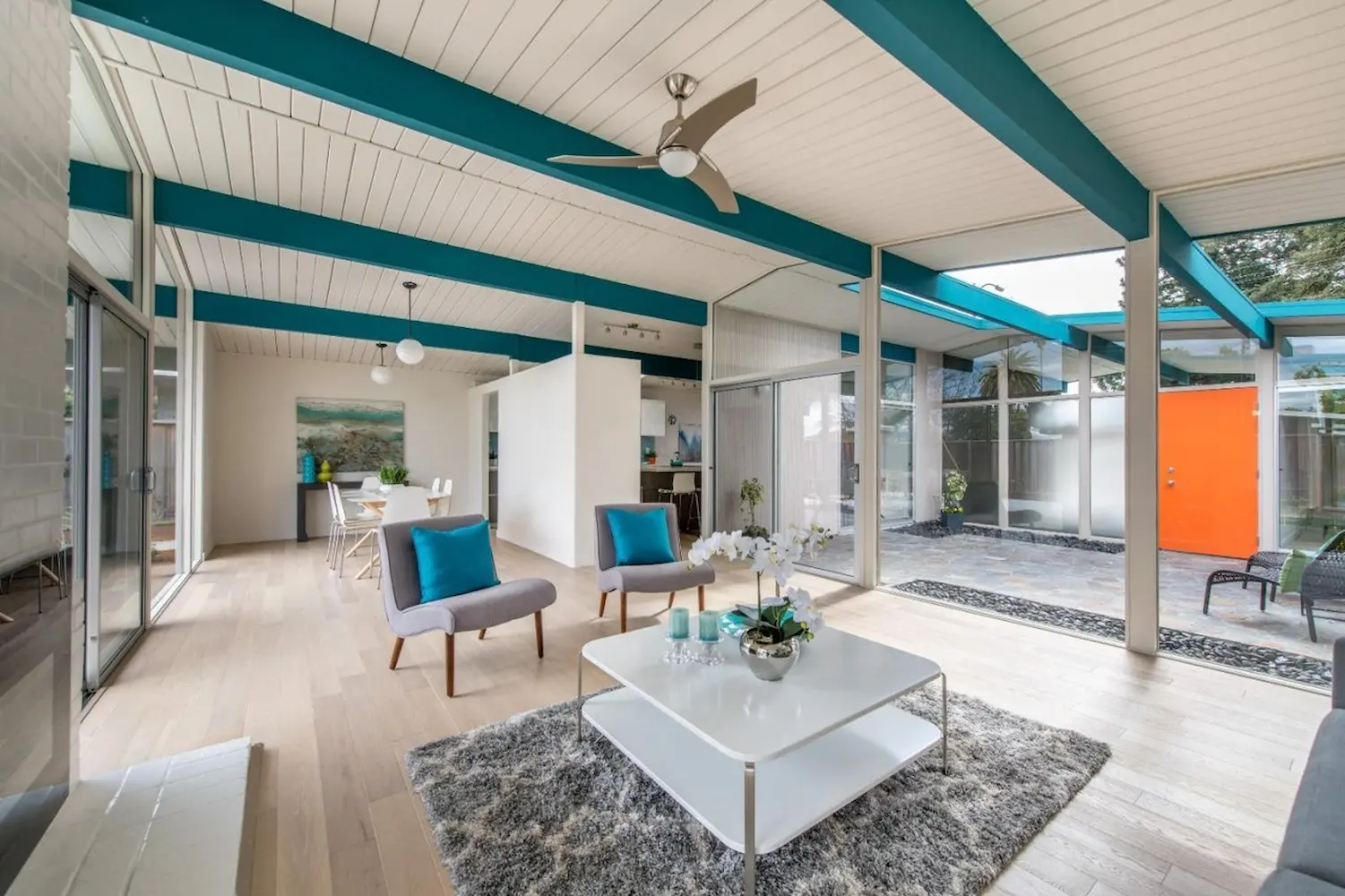 Modern open living room with teal ceiling beams, mid-century furniture, and large sliding glass doors leading to an outdoor courtyard