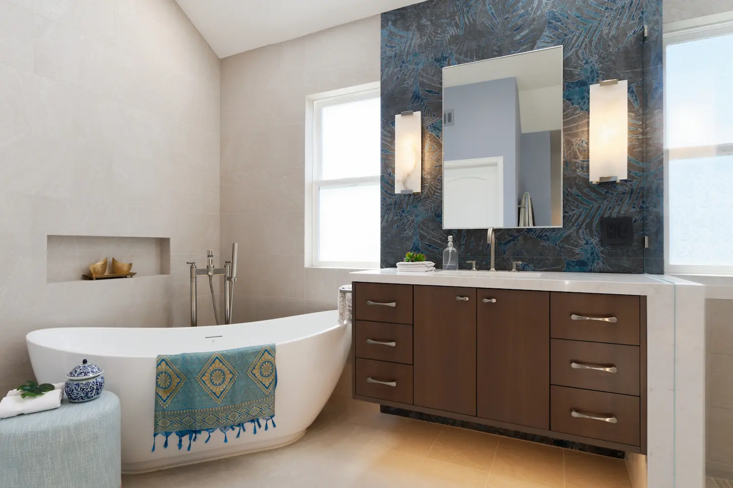 Modern bathroom design featuring a freestanding bathtub, wood vanity with white countertop, blue accent wall with geometric patterns, and wall-mounted lighting fixtures