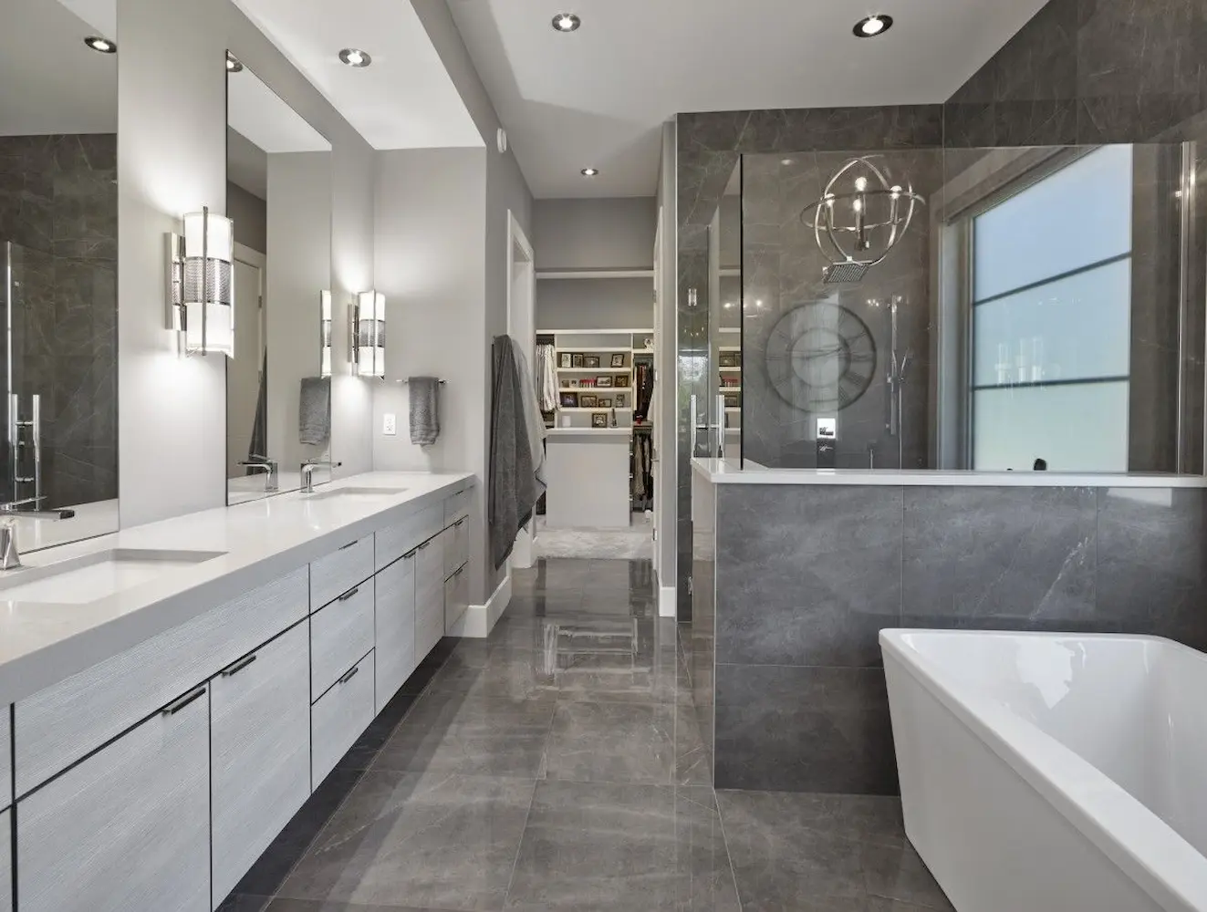 Luxury bathroom design featuring a dual-sink vanity with white countertops, modern lighting fixtures, large gray tiled walls, a freestanding bathtub, and a glass-enclosed walk-in shower