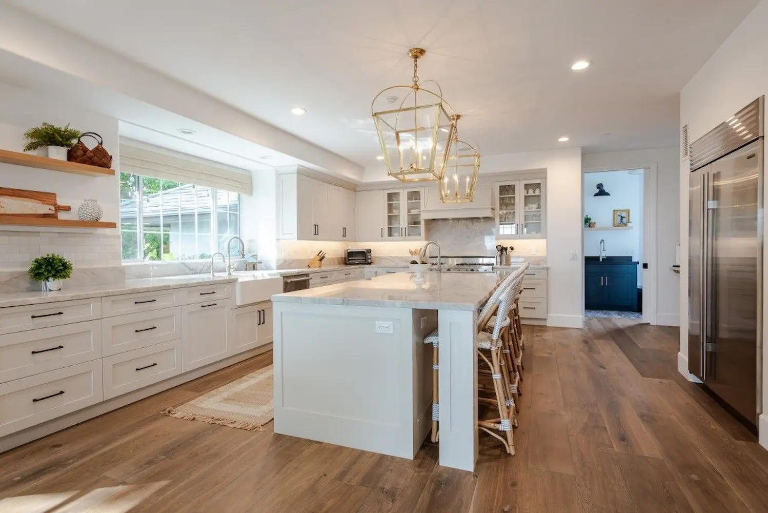 A spacious white kitchen with a large marble island, gold pendant lighting, open shelving, and farmhouse-style decor